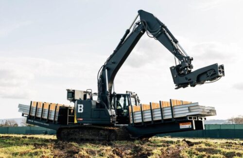 A machine resembling a backhoe has picks up a steel beam from a shelf built onto its side.